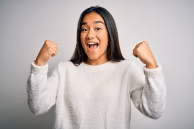 Genç ve güzel Asyalı kadın beyaz arka plan üzerinde sıradan bir süveter giyerek şaşkınlık ve şaşkınlık içinde kollarını kaldırarak ve gözlerini açarak başarıyı kutluyor. Kazanan konsept.