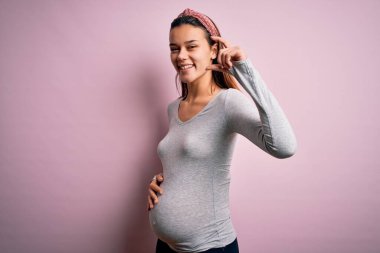 Genç, güzel bir kız hamile, pembe arka planda bebek bekliyor. Gülümsüyor ve el hareketleriyle küçük boy işaretler yapıyor. Parmakları ve kamerayla bakıyor. Ölçü kavramı.