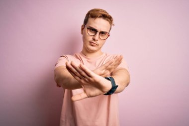 Genç, kızıl saçlı, sıradan bir tişört giyen, pembe arka planda duran, kollarını ve avuçlarını çaprazlayan, negatif işaret veren, kızgın yüzlü, yakışıklı bir adam.