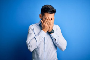 Zarif bir gömlek giyen yakışıklı bir adam, soyutlanmış mavi arka plan üzerinde duruyor, üzgün bir ifade var, elleri ile yüzünü kapatıyor ve ağlıyor. Depresyon kavramı.