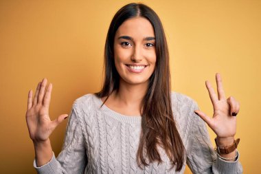 Sarı arka planda beyaz süveter giyen genç, güzel esmer kadın 8 numaralı parmaklarıyla işaret ederken kendinden emin ve mutlu gülümsüyor..