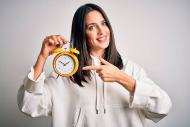 Mavi gözlü, çalar saati tutan genç bir kadın izole edilmiş beyaz arka planda duruyor. El ve parmağıyla işaret ederek çok mutlu.