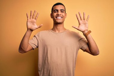Genç, yakışıklı, Afro-Amerikalı, günlük tişörtler giyen, sarı arka plan üzerinde duran ve parmaklarını on numarayı gösteren, kendinden emin ve mutlu gülümseyen bir adam..