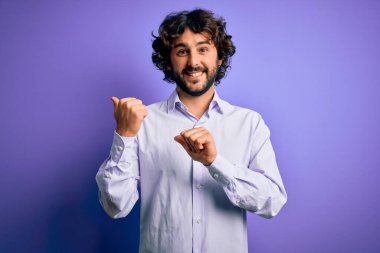 Mor arka planda duran sakallı, genç ve yakışıklı iş adamı. Arka tarafı işaret ediyor. El ve baş parmakları havada, kendine güvenen.