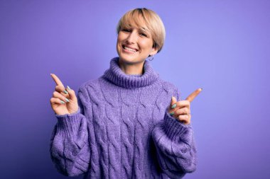 Kısa saçlı, mor arka planda balıkçı yaka kazak giyen, kendine güvenen, parmakları farklı yönleri gösteren genç sarışın kadın. Reklam için boşluk kopyala