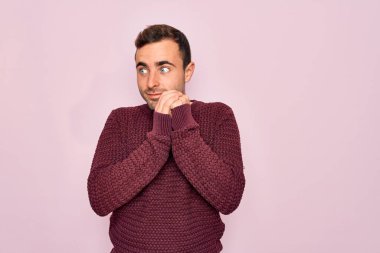 Mavi gözlü, sıradan bir süveter giyen, pembe arka planda duran, gergin ve heyecanlı, elleri çenesinde yan tarafa bakan genç yakışıklı bir adam.