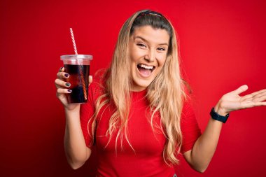 Genç güzel sarışın kadın kırmızı arka planda saman kullanarak kola köpüklü içecek içiyor. Çok mutlu ve heyecanlı, kazanan ifade zaferi büyük bir gülümsemeyle kutluyor ve ellerini kaldırıyor.