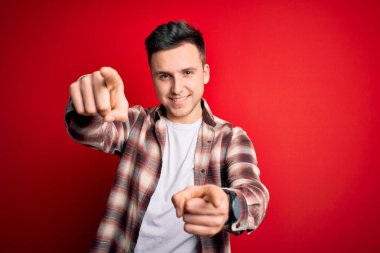 Kırmızı izole edilmiş arka plan üzerine gündelik modern tişört giyen genç beyaz adam seni ve kamerayı parmaklarıyla göstererek pozitif ve neşeli gülümsüyor.