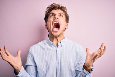 Sarışın, kıvırcık saçlı, beyaz arka planda çizgili tişört giyen çılgın ve sinirli, agresif bir ifade ve kollarla bağıran genç bir adam. Hayal kırıklığı konsepti.