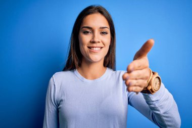 Young beautiful brunette woman wearing casual sweater standing over blue background smiling friendly offering handshake as greeting and welcoming. Successful business.