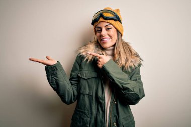 Genç esmer kayakçı kadın kar kıyafetleri ve kayak gözlükleri takmış beyaz arka planda hayrete düşmüş ve kameraya gülümsüyor..