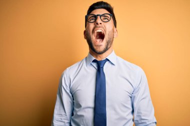 Kravat ve gözlük takan genç yakışıklı işadamı sarı arka planda kızgın ve öfkeli bir şekilde bağırıyor. Öfkeyle bağırıyor. Öfke ve agresif kavram.