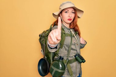 Sırt çantası ve sarı arka planda şapka takan kızıl saçlı genç bir kadın. Parmağını kaldırmış ve kızgın bir ifadeyle işaret ediyor.