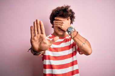 Çizgili sıradan tişörtler giyen genç yakışıklı adam izole edilmiş pembe arka plan üzerinde duruyor gözleri ellerle kapıyor ve üzgün ve korku ifadeleriyle jest yapıyor. Utanç verici ve olumsuz bir kavram..