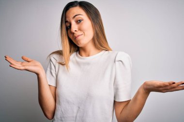 Genç, sarışın, mavi gözlü, beyaz arka planda sıradan tişörtler giyen, elleri ve kolları havada, kafası karışık genç bir kadın. Şüphe kavramı.