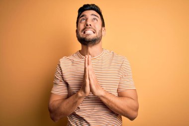 Sakallı genç yakışıklı bir adam sarı arka planda sıradan bir tişört giyiyor. Elleri bağlı yalvarıyor ve dua ediyor. Yüzünde umut ifadesi var. Yalvarıyorum..