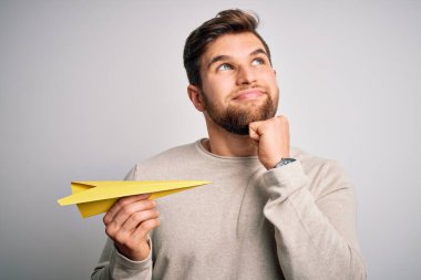 Sarışın, sakallı, mavi gözlü beyaz arka planda kağıt uçak tutan, soru soran, kafası karışık genç bir adam.