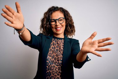 Orta yaşlı esmer iş kadını gözlüklü, izole edilmiş beyaz arka planda durup kameraya sarılmak için gülümseyerek bakıyor. Mutluluğu kucaklayan neşeli bir ifade..