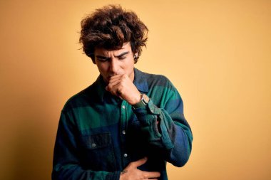 İzole edilmiş sarı arka plan üzerinde dikilen genç ve yakışıklı bir adam kendini iyi hissetmiyor ve soğuk algınlığı veya bronşitin belirtisi olarak öksürüyor. Sağlık hizmeti kavramı.