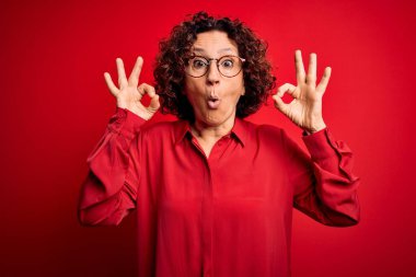Orta yaşlı güzel kıvırcık saçlı kadın kırmızı arka planda günlük gömlek ve gözlük takıyor. Şaşırmış ve şaşırmış görünüyor. Çılgın ifade