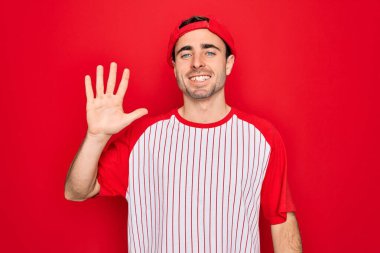 Mavi gözlü, çizgili beysbol tişörtü ve şapkası giyen genç yakışıklı sporcu beş numaralı parmaklarını gösterirken kendinden emin ve mutlu bir şekilde gülümsüyor..