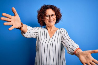 Orta yaşlı güzel kıvırcık saçlı kadın izole arka planda sıradan çizgili tişört giyiyor ve kameraya sarılıp gülümsüyor. Mutluluğu kucaklayan neşeli bir ifade..