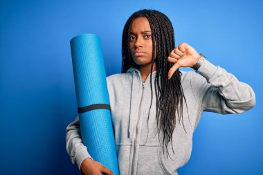 Genç Afro-Amerikan fitness kadını yoga minderini izole edilmiş mavi arka planda tutuyordu. Kızgın bir yüzü vardı. Eksi işareti baş parmakları aşağıda, reddedilme konsepti vardı.