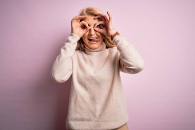 Orta yaşlı güzel sarışın kadın pembe arka planda balıkçı yaka kazak giyiyor. Dürbün gibi güzel hareketler yapıyor. Gözleri parmaklarından bakıyor. Çılgın ifade.