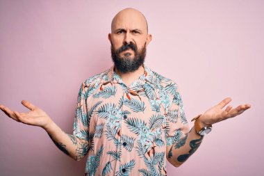 Sakallı ve dövmeli yakışıklı, pembe arka planda sıradan bir çiçek gömleği giyen, kolları açık ve kafası karışık, hiçbir fikri olmayan bir adam..