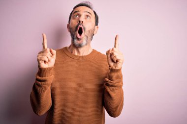 Orta yaşlı, beyaz, günlük kahverengi süveter giyen, pembe arka planda dikilen, şaşkın, şaşkın, parmaklarıyla işaret eden ve kollarını kaldıran bir adam..
