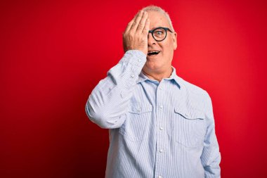 Orta yaşlı, yakışıklı, beyaz bir adam kırmızı arka planda günlük çizgili gömlek ve gözlük takıyor. Bir gözünü eliyle kapatıyor. Yüzünde kendine güvenen bir gülümseme ve sürpriz bir duygu var..