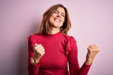 Genç, güzel, esmer bir kadın pembe arka planda sıradan bir süveter giyiyor. Çok mutlu ve heyecanlı. Kolları havada, gülümseyerek ve başarı için çığlık atarak kazanan jestler yapıyor. Kutlama konsepti.