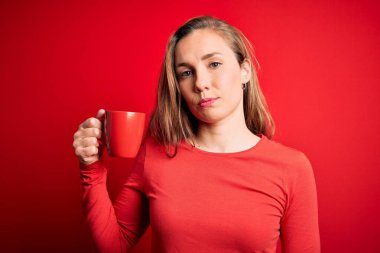Genç, sarışın bir kadın, izole edilmiş kırmızı arka planda duran bir fincan kahve içiyor. Yüzünde kendine güvenen bir ifade var ve ciddi düşünüyor.