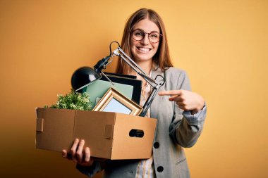 Genç ve güzel kızıl saçlı iş kadını sarı arka planda karton kutuyu tutarak ateş etti. Çok mutlu bir şekilde eli ve parmağı işaret etti.
