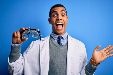 Yakışıklı afro-amerikan optik adam mavi arka planda optometri gözlüklerini tutuyor çok mutlu ve heyecanlı, kazanan yüz ifadesi zaferi kutluyor büyük bir gülümsemeyle ve el kaldırarak