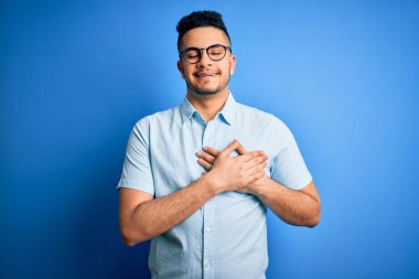 Genç, yakışıklı bir adam, gözlüklü, sıradan bir tişört ve gözlüklü, mavi arka planda, elleri göğsünde, gözleri kapalı ve yüzünde minnet dolu bir jest ile gülümsüyor. Sağlık kavramı.