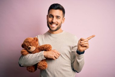 Genç yakışıklı bir adam, ayıcığı soyutlanmış pembe arka planda tutarken çok mutlu bir şekilde elini ve parmağını kenarı işaret ediyor.