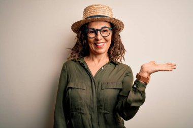 Gözlüklü ve şapkalı orta yaşlı esmer kadın izole edilmiş beyaz arka planda duruyor neşeli bir şekilde gülümsüyor ve avuç içi kameraya bakıyor..