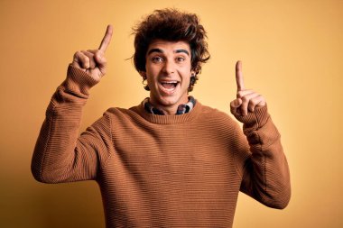 Sarı arka planda rahat bir gömlek ve süveter giyen genç yakışıklı adam şaşırmış, şaşırmış ve parmaklarıyla işaret ederek kollarını kaldırmış..