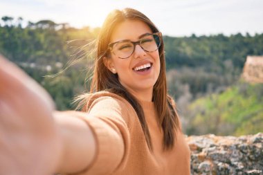 Genç, güzel, esmer, gözlüklü, mutlu ve kendinden emin bir kadın. Yüzünde gülümsemeyle şehrin caddesindeki kameranın önünde selfie çekerken.