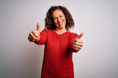 Orta yaşlı esmer kadın izole edilmiş beyaz arka planda dikilip olumlu bir jest yapmayı onaylıyor, gülümsüyor ve başarı için mutlu. Kazanan jest.