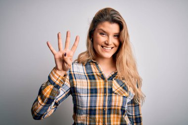 İzole edilmiş beyaz arka plan üzerinde duran ve dört numaralı parmaklarıyla işaret eden, kendinden emin ve mutlu gülümseyen genç sarışın kadın..