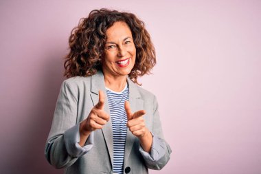 Soyutlanmış pembe arka planda zarif bir ceket giyen orta yaşlı güzel iş kadını mutlu ve komik yüzlü parmaklarını kameraya doğrultuyor. İyi enerji ve titreşimler.