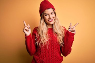 Genç, sarışın, sıradan bir kazak giyen, beyaz arka planda yün bir şapka takan, kendine güvenen, parmakları farklı yönleri gösteren güzel bir kadın. Reklam için boşluk kopyala