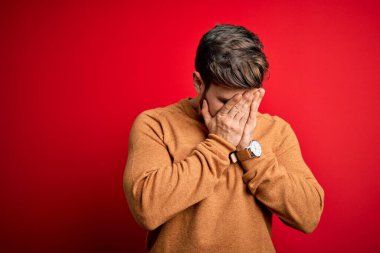 Sakallı ve mavi gözlü genç bir adam kırmızı arka planda sıradan bir süveter giyiyor ve ağlarken suratını ellerle kapatıyor. Depresyon kavramı.