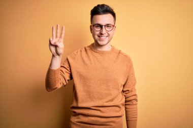 Sarı arka planda gözlüklü, beyaz, yakışıklı, gözlüklü ve sıradan bir süveter giyen genç adam üç numarayı gösterirken, kendinden emin ve mutlu bir şekilde gülümsüyor..