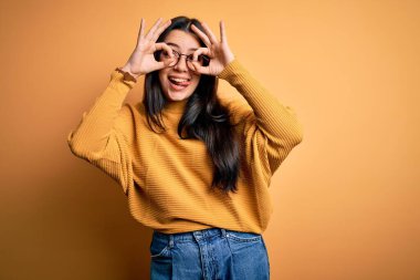 Genç esmer kadın gözlük takıyor ve sarı izole edilmiş arka plan üzerine gündelik süveter giyiyor. Dürbün gibi jest yapıyor, gözleri parmaklarından bakıyor. Çılgın ifade.