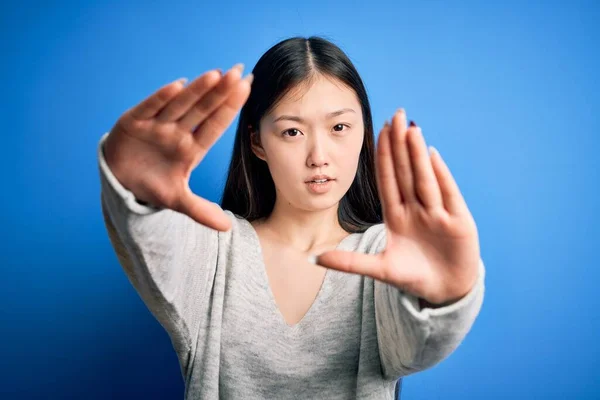 Young beautiful asian woman wearing casual sweater standing over blue isolated background doing frame using hands palms and fingers, camera perspective