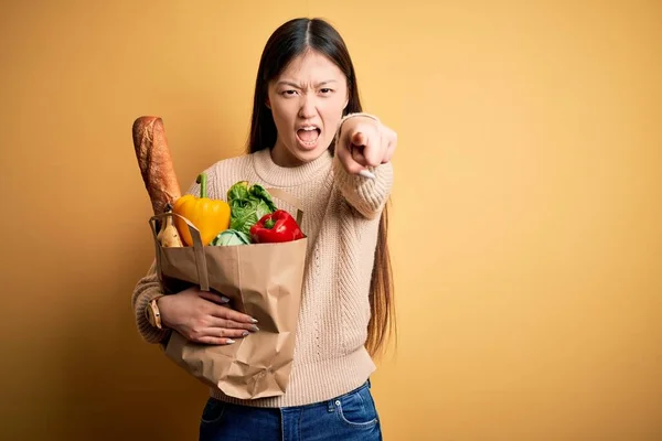 年轻的亚洲女人拿着一袋袋新的健康食品放在黄色孤立的背景上 带着不满和沮丧的心情看着摄像机 对你怒气冲冲 — 图库照片