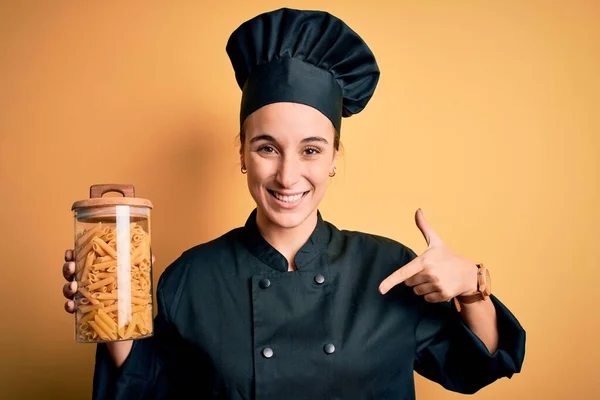 年轻美丽的厨师女士身穿厨师制服 手里拿着盛有意大利面通心粉的罐子 满脸惊讶 手指指向自己 — 图库照片